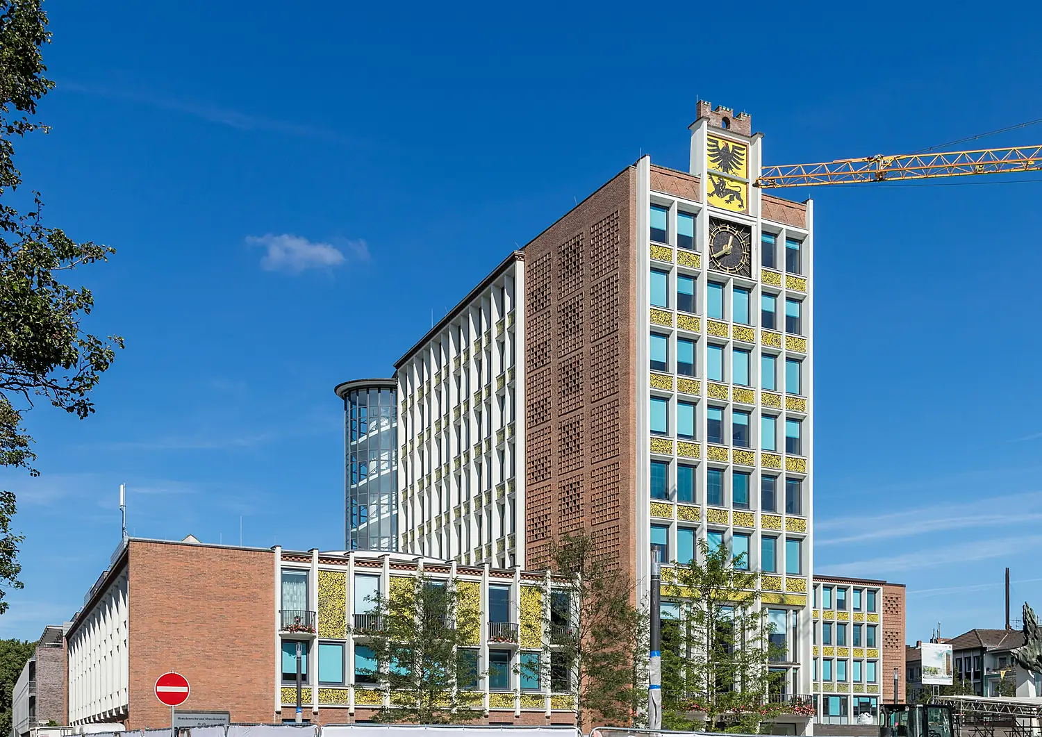 Das Rathaus Düren mit Klinkerfassade, Uhr und Wappen am Turmaufsatz, davor Bäume, unter blauem Himmel