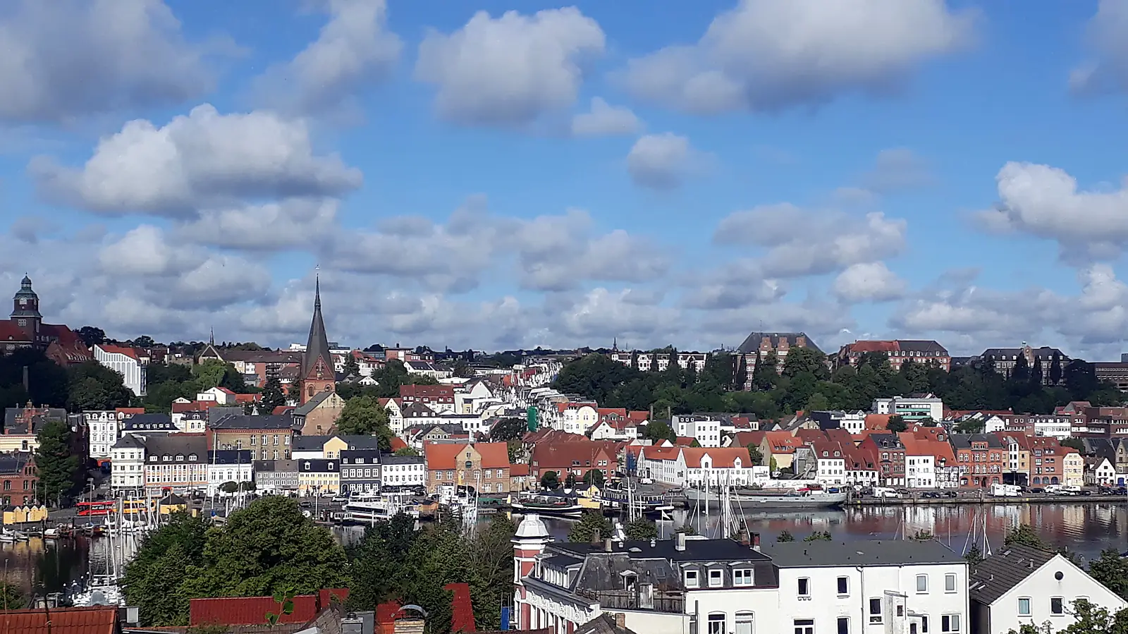 Blick über den Hafen auf die Flensburger Altstadt mit roten Dächern und einem Kirchturm unter Wolkenhimmel