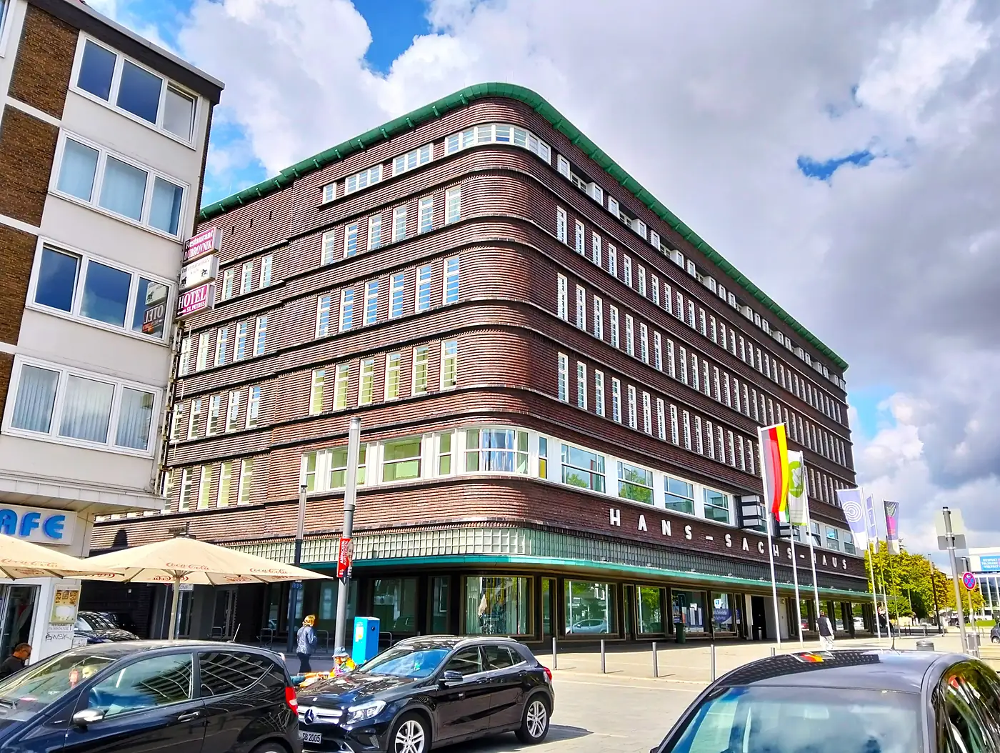 Hans-Sachs-Haus in Gelsenkirchen, ein Backsteinbau mit abgerundeter Ecke und grünem Dachrand unter Wolkenhimmel