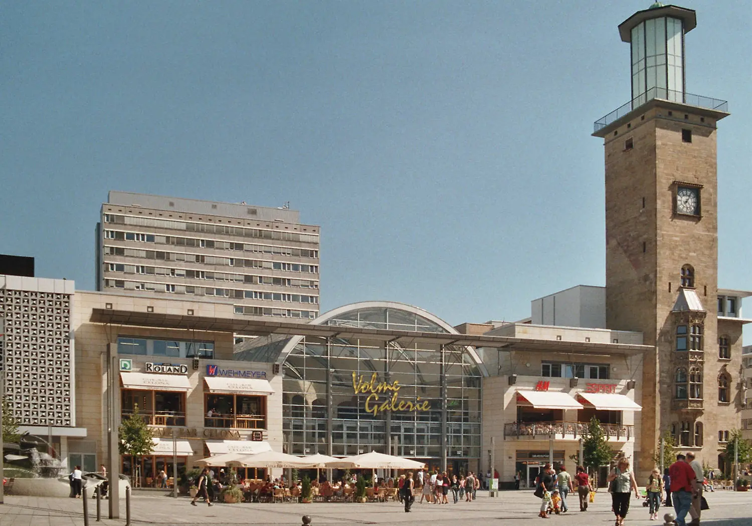 Turm mit Uhr und gewölbtem Glasdach einer Passage am Rathausplatz in Hagen, davor Passanten und Sonnenschirme
