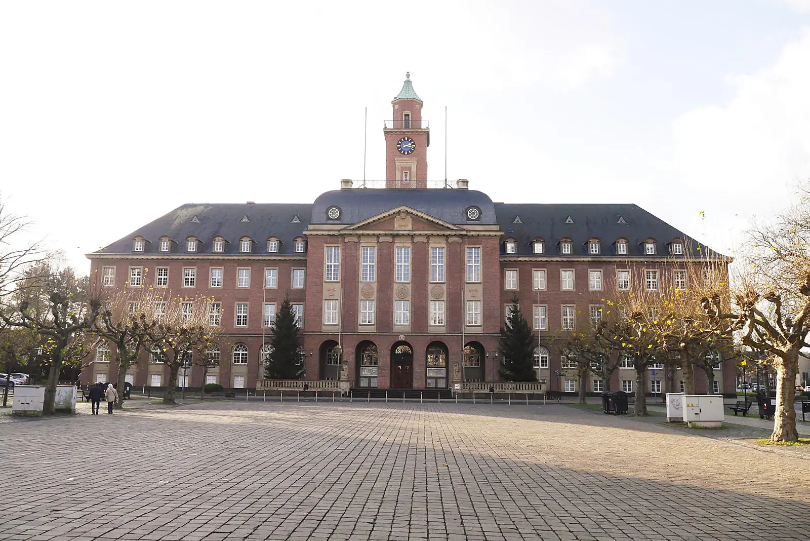 Das Rathaus von Herne, ein symmetrischer Backsteinbau mit Uhrturm, davor ein gepflasterter Platz mit Bäumen