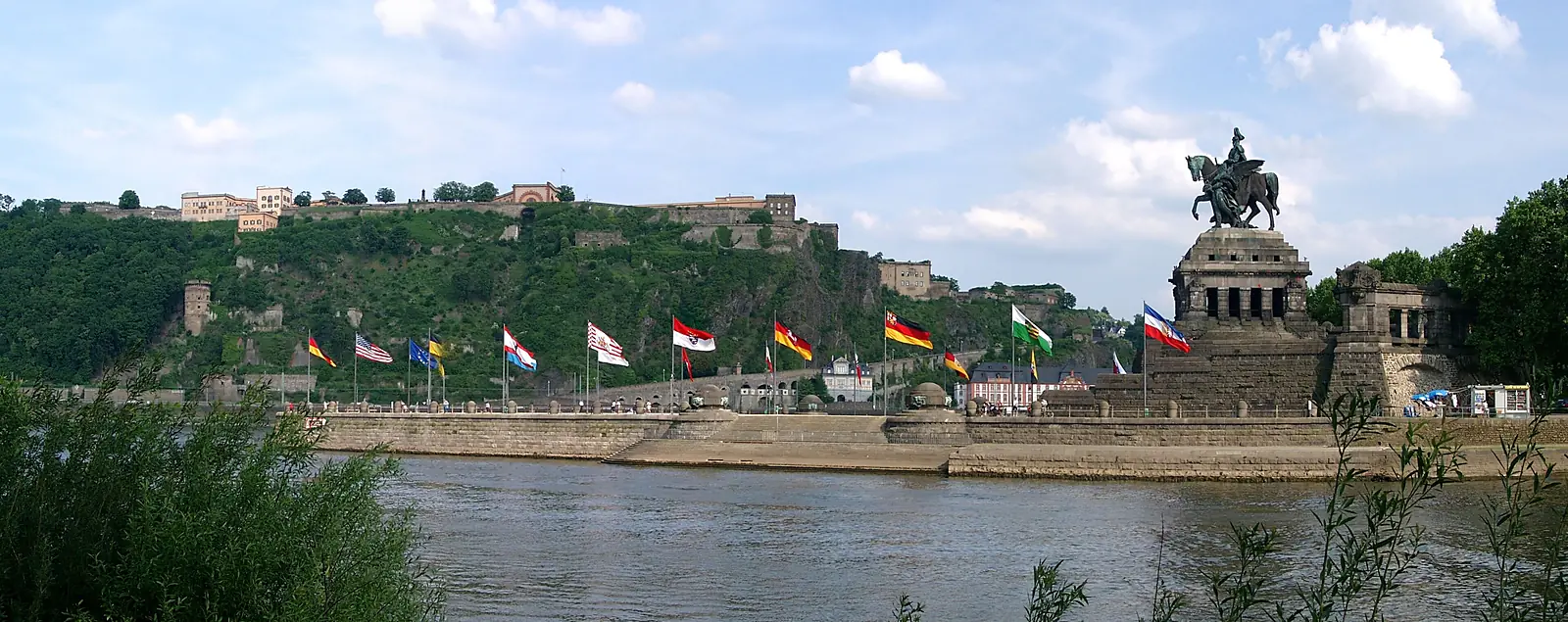 Das Deutsche Eck in Koblenz mit dem Reiterdenkmal und Flaggen am Ufer, dahinter ein bewaldeter Hang mit Gebäuden