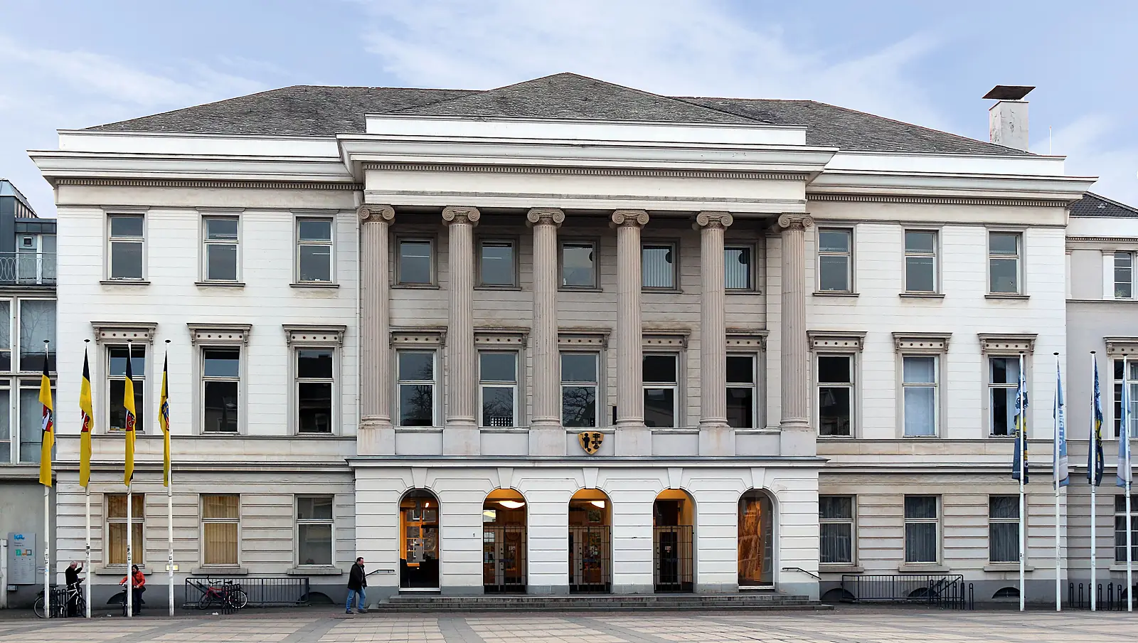 Rathaus Krefeld, weiße klassizistische Fassade mit sechs Säulen und fünf Rundbogen-Eingängen, davor Fahnen auf dem Platz