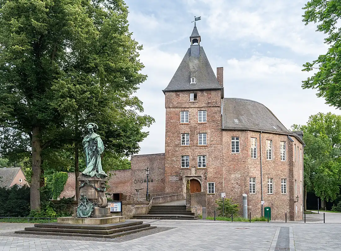 Das Schloss Moers, ein Backsteinbau mit spitzem Turm, davor eine Figur auf einem Sockel und alte Bäume
