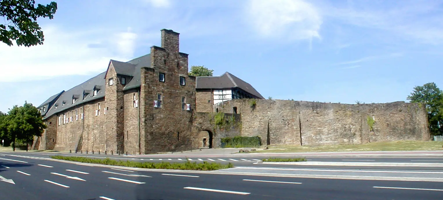 Schloss Broich in Mülheim an der Ruhr, eine Burg aus Naturstein mit Mauer, davor eine Straße unter blauem Himmel