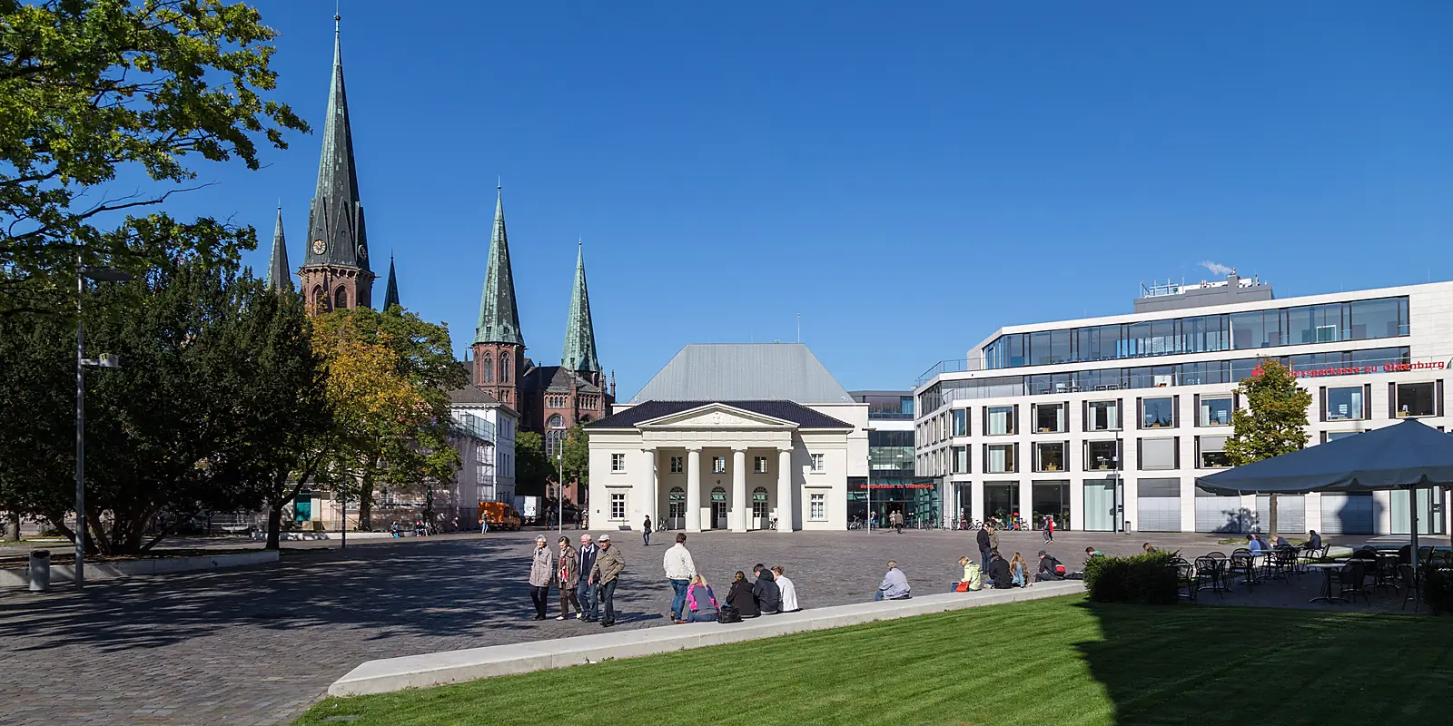 Schlossplatz in Oldenburg mit den Türmen der Lambertikirche, einem klassizistischen Bau und Passanten bei blauem Himmel