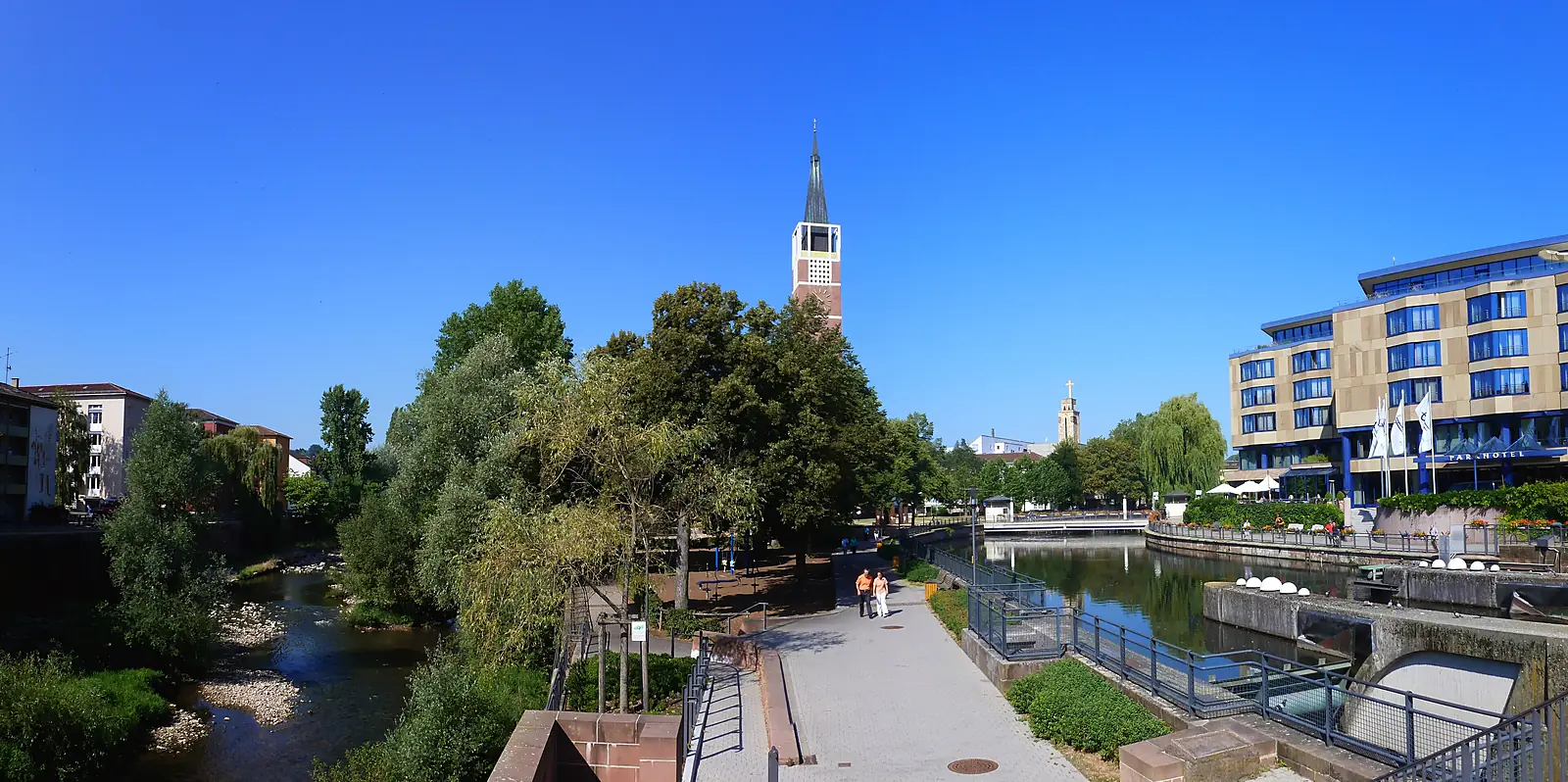 Fluss in Pforzheim mit Uferweg, Kirchturm im Hintergrund und Hotelgebäude rechts unter blauem Himmel