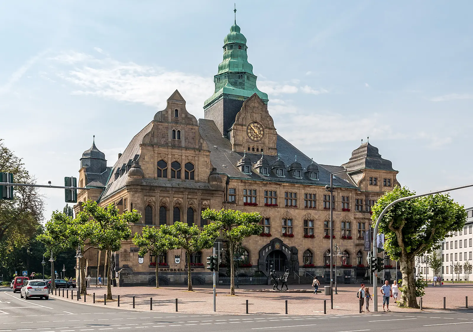 Das Rathaus Recklinghausen mit grünem Turm, Uhr und Sandsteinfassade, davor Bäume und ein offener Platz