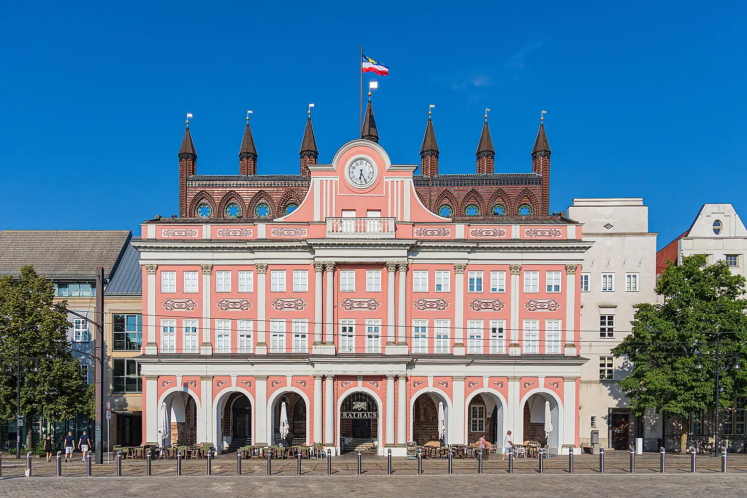 Rostocker Rathaus mit rosa Barockfassade, dahinter die Backsteingiebel mit spitzen Türmchen, blauer Himmel