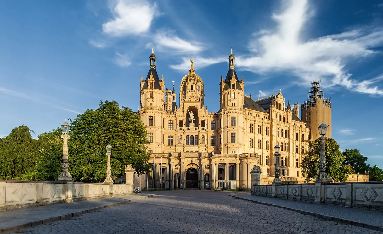 Schweriner Schloss im Abendlicht von der Brücke aus gesehen, mit Türmen und goldener Kuppel unter blauem Himmel