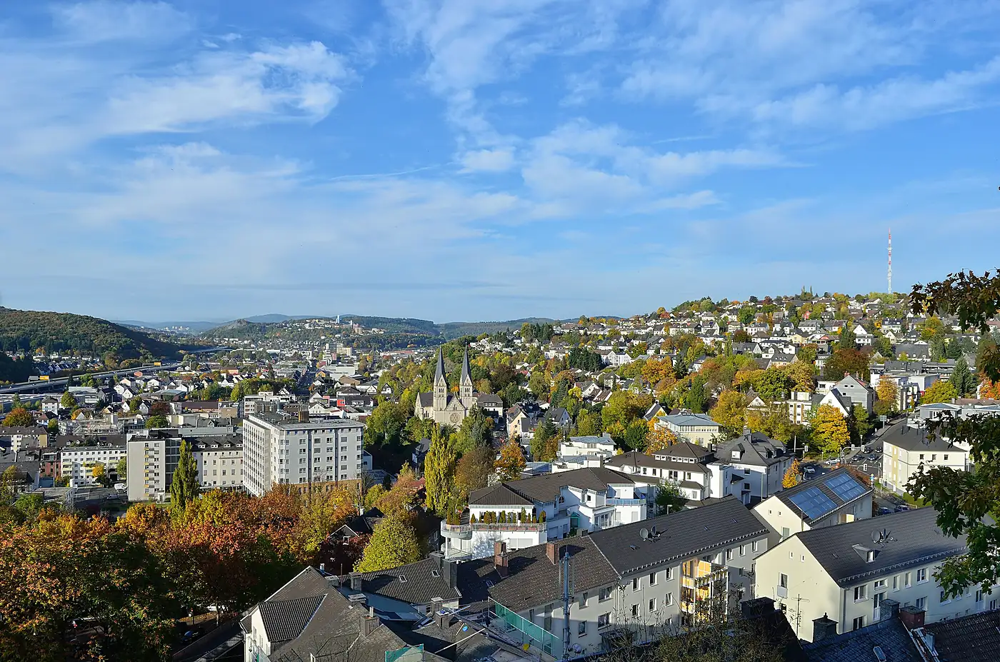 Blick über Siegen im Herbst, Wohnhäuser am Hang und eine Kirche mit zwei Türmen, im Hintergrund bewaldete Höhen